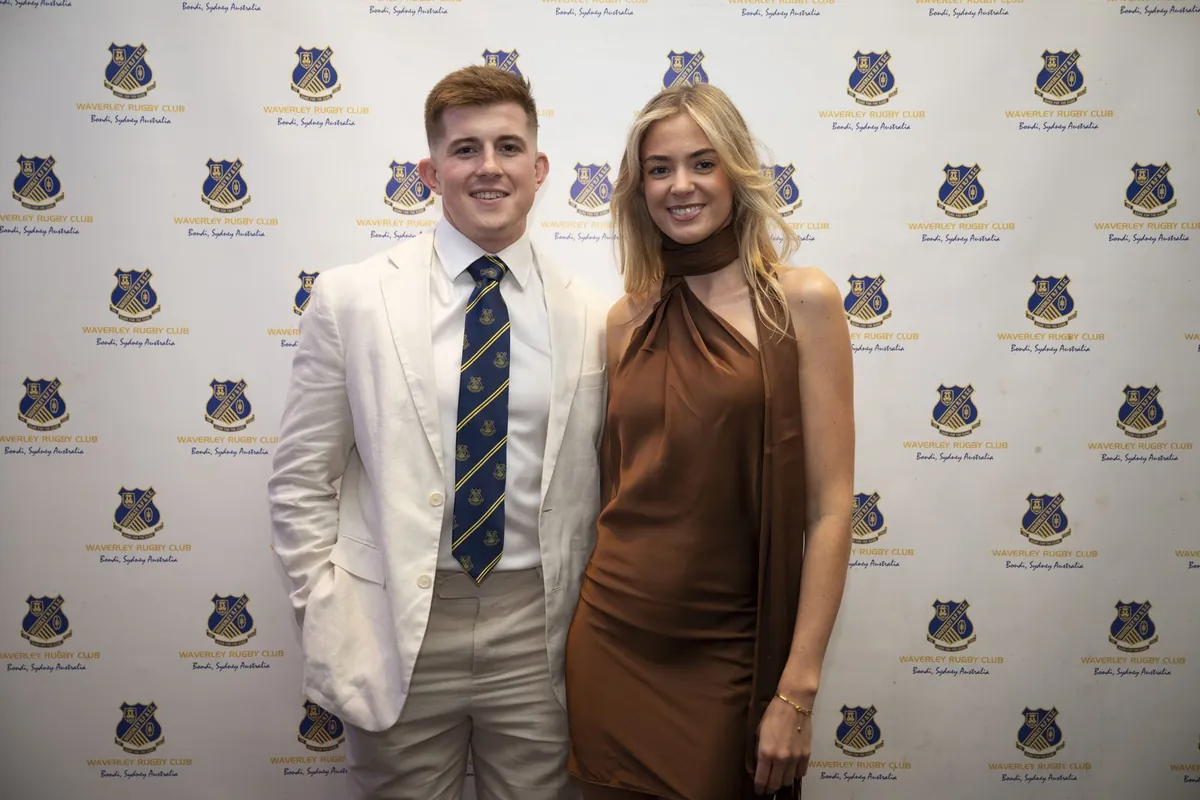 A couple in formalwear — a man in a white blazer with the club tie and a woman in a brown satin dress — posing in front of the Waverley Rugby backdrop.
