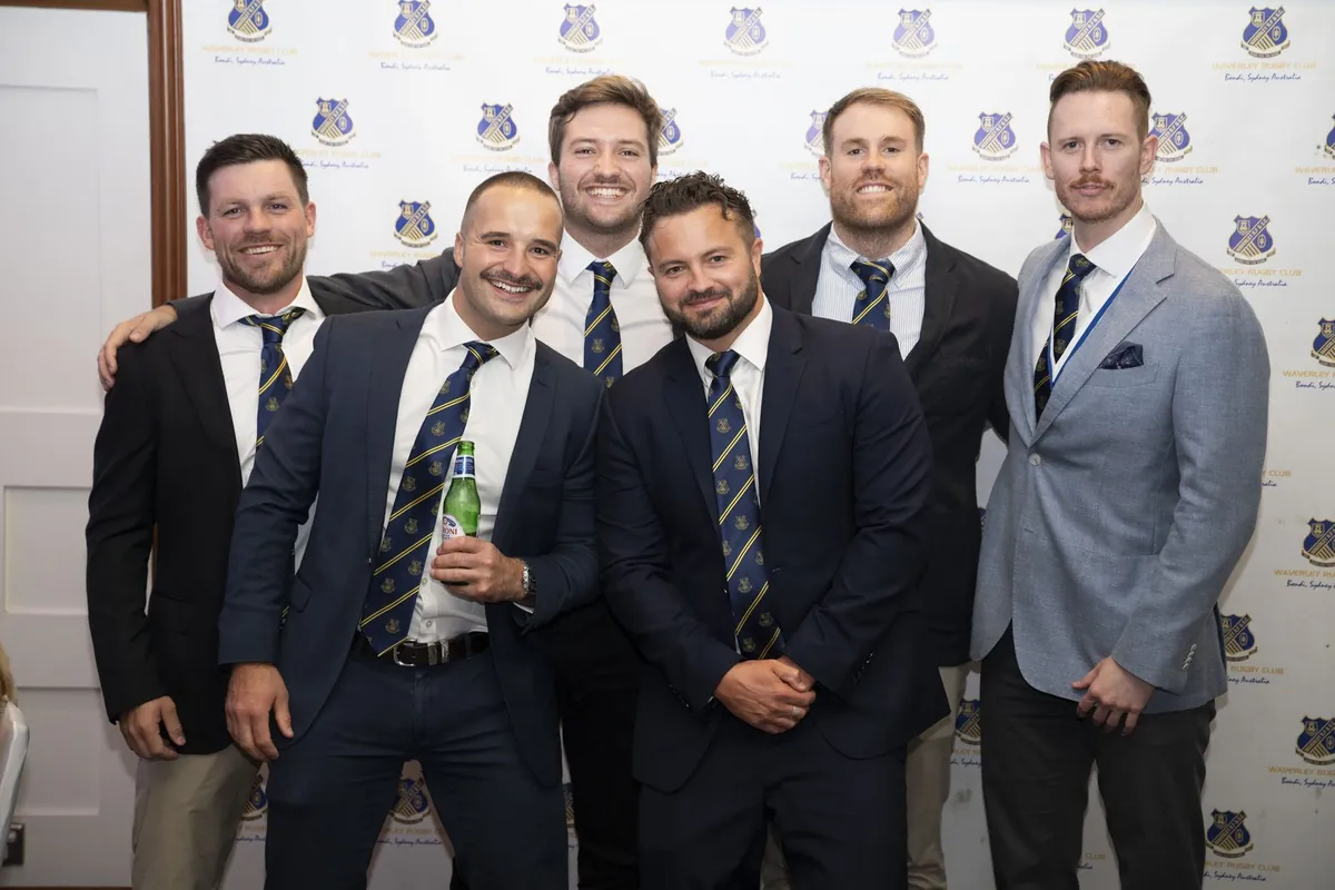 Six clubmates in blazers and club ties posing together in front of the club photo wall.
